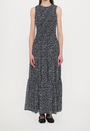 Maxi dress in black fabric with small blue floral pattern. Sleeveless design with gathered bodice and tiered skirt. Worn with black flat shoes.