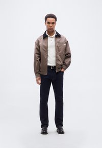 Καφέ ελαφρύ τζάκετ με μαύρο γιακά, φορεμένο πάνω από λευκή πόλο μπλούζα· συνδυασμένο με σκούρο τζιν και μαύρα παπούτσια.