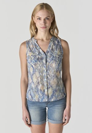 Mujer rubia con grandes pendientes de aro, blusa sin mangas con estampado y botones, y shorts de mezclilla remangados, de pie contra un fondo liso.