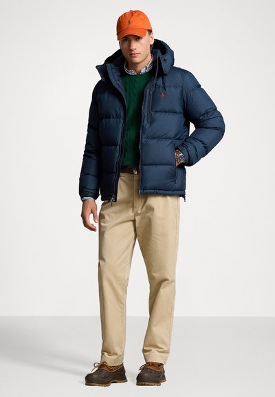 Giacca puffer blu navy con cappuccio staccabile, indossata sopra un maglione verde a trecce. Pantaloni chino beige e stivali impermeabili marroni. Un cappello arancione completa il look.