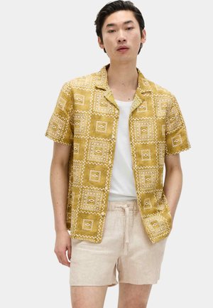 Joven con camisa de manga corta de color mostaza con patrón sobre camiseta blanca y pantalones cortos beige con cordón, posando frente a un fondo liso.