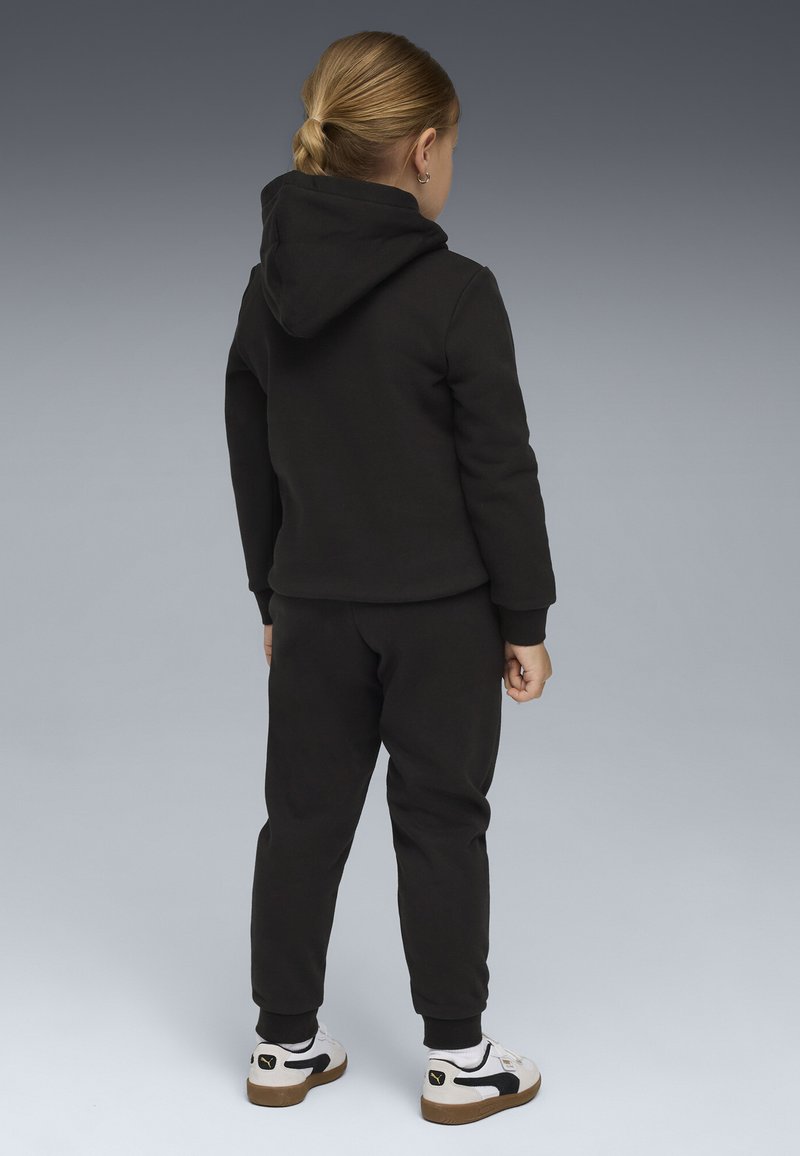 Schwarzes Baumwoll-Hoodie- und Jogginganzug-Set, mit Kapuze, gerippten Bündchen und tailliertem Design. Kombiniert mit schwarz-weißen Sneakers.