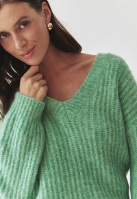 Pull en tricot vert menthe avec un col en V, texture côtelée et coupe ample. Il a des manches longues et un tissu doux et pelucheux.