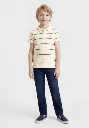 Blond chłopiec stojący, ubrany w jasnożółtą koszulkę polo w paski, ciemnoniebieskie jeansy i granatowe wsuwane buty na tle jednolitego tła.