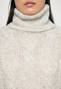 Pull à col roulé en tricot torsadé gris clair avec une texture douce et duveteuse. Présente des accents côtelés et un col haut. Vue rapprochée de l'encolure et du motif.