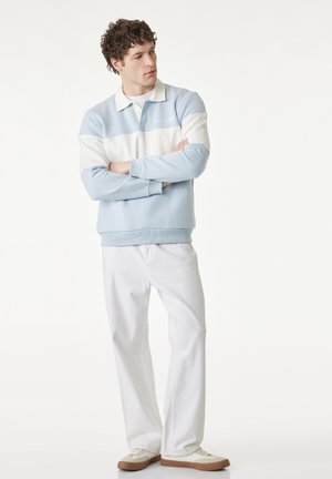 Mężczyzna stojący z założonymi ramionami, ubrany w jasnoniebiesko-białą koszulkę polo z długim rękawem, białe spodnie i beżowe trampki na białym tle.