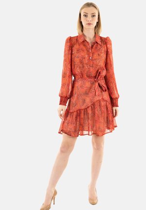 Vestido de estampado floral rojo con mangas largas, cuello y cinturilla. Tejido transparente con dobladillo en capas con volantes. Llevado con tacones nude.