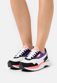 Pieds portant des baskets Puma blanches, noires, violettes et corail avec des lacets blancs et des touches vertes sur un fond blanc.