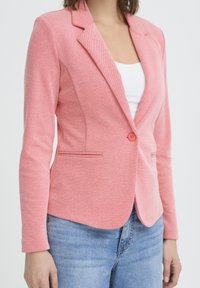Blazer rose en tissu texturé, avec une coupe cintrée, des revers crantés et une fermeture à un bouton. Poches sur le devant.