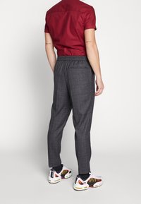 Man met grijs geblokte broek, zwarte sokken, witte sneakers met rode en gele details, en een kortemouw rood shirt, staande met de rug naar de kijker.