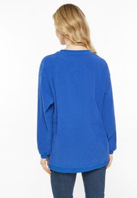 Kék, bordázott pulóver hosszú ujjal, kerek nyakkivágással és laza szabással. Az alja a csípőn túl ér, puha anyagból készült.