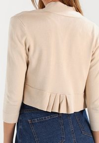 Bežový krátký kardigan s skládacím zadním detailem, nošený přes džínovou sukni, zobrazený zboku na osobě s dlouhými vlasy.