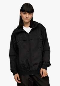 Giacca nera con cerniera, cappuccio e due tasche anteriori. Realizzata in tessuto lucido e strutturato con polsini elasticizzati. Design semplice e moderno.