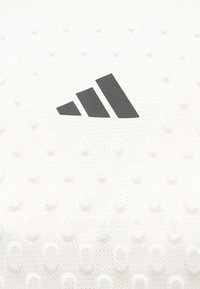 Logo Adidas noir à trois bandes centré sur un tissu blanc texturé avec de petits points en relief et un motif en maille respirante.