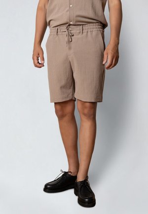 Mand iført teksturerede beige shorts med elastisk taljebånd og snøre, kombineret med sorte snøresko, stående mod en ensfarvet baggrund.