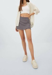 Grijze mini rok met een lichte asymmetrische zoom, gecombineerd met een witte geribbelde crop top en een crèmekleurig gestreept oversized shirt. Stevige beige instappers.