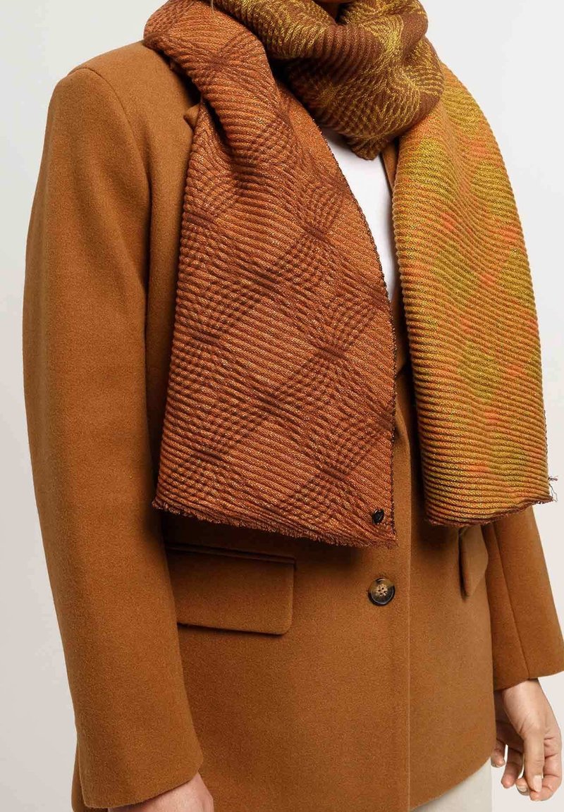 Manteau en laine marron avec bouton, porté sous une écharpe épaisse texturée aux tons orange et vert, enroulée autour du cou.