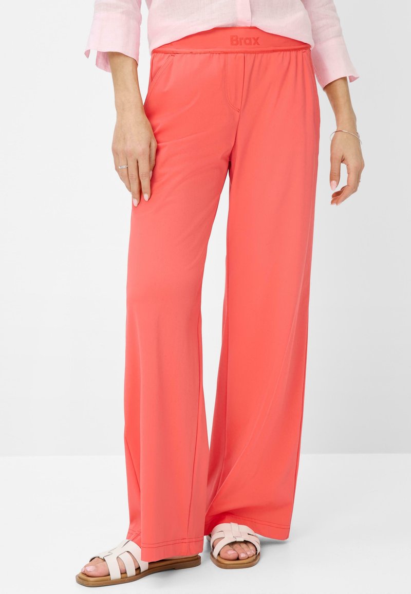 Femme portant un pantalon corail à jambes larges, une chemise rose clair, des sandales blanches et des bagues et un bracelet en argent.