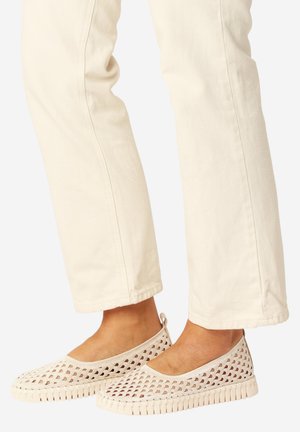 Voeten met beige geweven instapschoenen gecombineerd met off-white rechte broeken tegen een effen achtergrond.