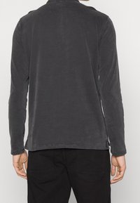 Halenka s dlouhým rukávem z tmavě šedé látky s hladkým povrchem. Obsahuje kulatý výstřih a postranní rozparky na spodním lemu pro snadnější pohyb.