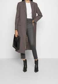 Blazer largo gris claro con hombros estructurados, sobre una blusa blanca y jeans gris oscuro. Botines negros y un bolso negro completan el look.