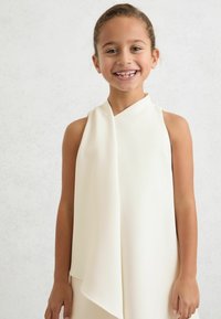 Ujjatlan, krémszínű ruha drapériás kivitelben, mely sima, könnyű anyagból készült és laza szabású.