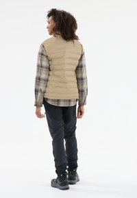 Beige pufferväst, rutmönstrad långärmad skjorta, svarta byxor och grå sneakers. Västen har en hög krage och ett kviltat mönster.
