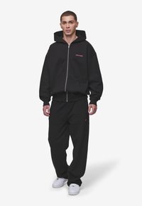 Black zip-up hoodie with a large hood and red logo on the chest, paired with loose black pants and white sneakers. Soft fabric texture.