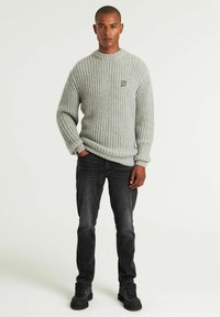 Hellgrauer gestrickter Pullover mit rippenstruktur und Rundhalsausschnitt, kombiniert mit dunklen Jeans und schwarzen Stiefeln. Besitzt ein kleines Logo auf der Brust.