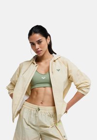 Mladá žena s tmavými vlasmi má na sebe béžovú vetrovku a šortky cez zelenú športovú podprsenku, pózuje s rukami v bok na bielom pozadí.