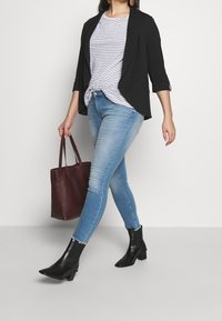 Veste noire sur une chemise à rayures avec un nœud à l'avant, associée à un jean skinny bleu clair et des bottines noires. Sac fourre-tout bordeaux à la main.