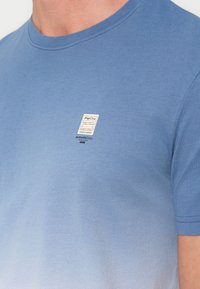 Άνδρας που φοράει μπλε μπλουζάκι με διαβαθμισμένο χρώμα και κοντά μανίκια, με μικρό ορθογώνιο λογότυπο "Pier One" στο αριστερό μέρος του στήθους.
