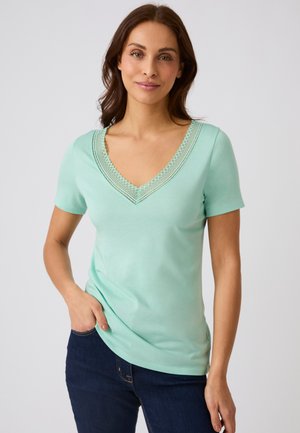 Vrouw met bruin haar draagt een lichtgroen T-shirt met korte mouwen en V-hals met kantafwerking, gecombineerd met donkerblauwe jeans, staand tegen een effen achtergrond.