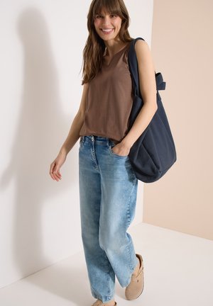 Lächelnde Frau mit braunem ärmellosem Top, hellblauen Jeans, beigen Slip-On-Schuhen, die eine große dunkelblaue Umhängetasche auf der Schulter trägt.