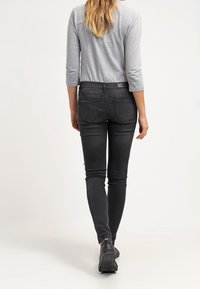 Jeans negros ajustados con cintura alta, bolsillos traseros y textura lisa, combinados con una camiseta gris de manga tres cuartos y zapatos negros.