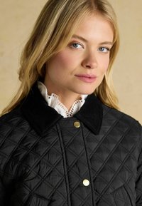 Jachetă neagră din material matlasat cu guler din catifea, butoane aurii cu capscă și un model texturat în diamant. Un guler alb cu volane este vizibil dedesubt.