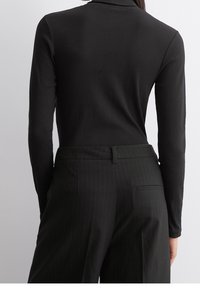 Schwarzes Langarmoberteil mit Rippstruktur, kombiniert mit dunklen Steifenhosentrousern, die über eine Gesäßtasche und Gürtelschlaufen verfügen.