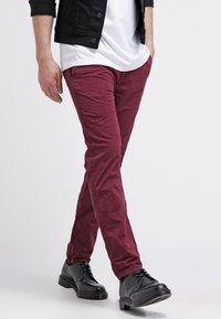 Pantalon bordeaux, ajusté, avec une texture froissée, associé à des chaussures noires à lacets et un t-shirt blanc sous une veste noire.