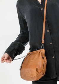 Borsa a tracolla in pelle marrone con finitura texturizzata, tracolla a catena e chiusura a scatto, indossata da una persona con una camicia scura con bottoni.