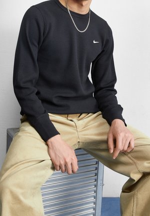 Homme assis sur un banc en métal portant un t-shirt Nike noir à manches longues, un pantalon beige et un collier chaîne argenté, mains posées sur les cuisses.
