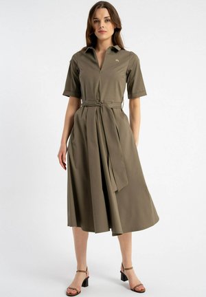 Kobieta stojąca, ubrana w oliwkową sukienkę midi z krótkimi rękawami, kołnierzykiem, przednim suwakiem, przewiązanym paskiem w talii oraz czarnymi sandałami na szerokim obcasie.