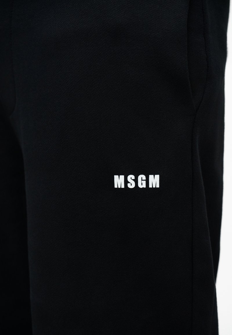 Zwarte stoffen broek met een klein wit MSGM-logo gedrukt op het bovenbeen vlakbij een zijzak.
