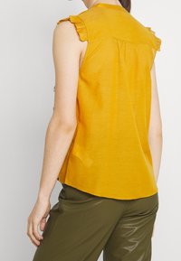 Hořčicově žlutý bezrukávový top s volánkovým detailem na rameni, hladkou texturou, zadním yoke a mírně zakřiveným lemem. Vzduchově spárovaný s olivově zelenými kalhotami.