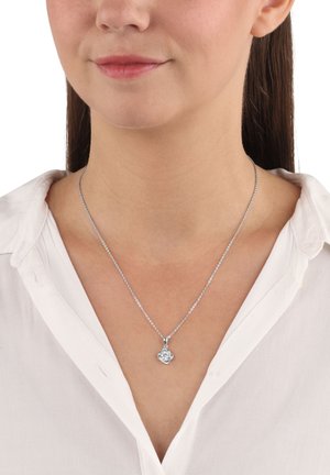 Collana d'argento con un pendente floreale, presenta una superficie texturizzata e una catena fine. Indossata con una camicia bianca con colletto.