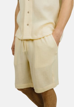 Mann iført lysebeige teksturerte shorts og matchende kortermet skjorte med knapper, med en hånd i lomma, mot hvit bakgrunn.
