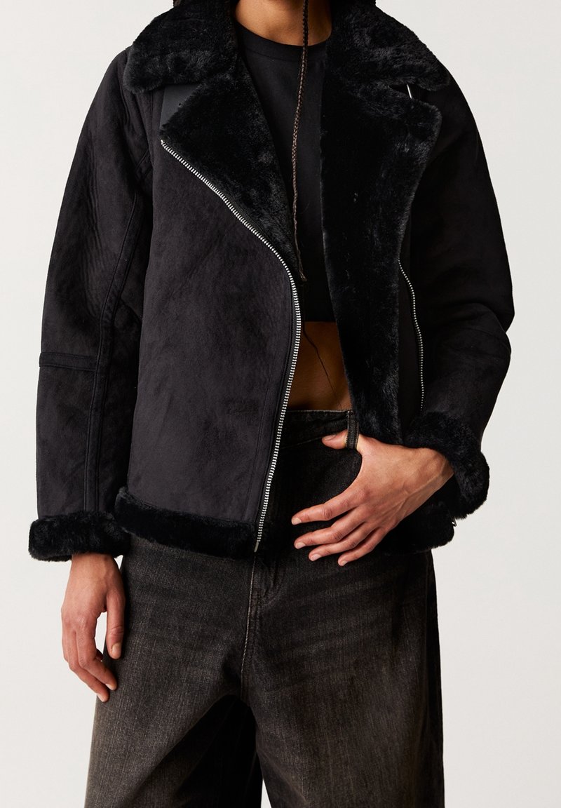 Personne vêtue d'une veste noire doublée de fausse fourrure, d'un haut court noir et d'un jean taille haute gris foncé, avec une main dans la poche.