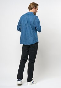 Langemouwig denim overhemd in blauw, met een kraag, rugpas en afgeronde zoom. Gecombineerd met zwarte broek en witte sneakers.