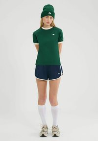 Zielona dopasowana koszula z kremowym wykończeniem, w zestawieniu z granatowymi szortami z białymi akcentami. Modelka nosi pasującą zieloną czapkę i białe skarpetki do kolan.