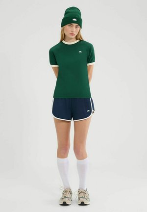 Groen getailleerd overhemd met crèmekleurige bies, gecombineerd met marineblauwe shorts met witte accenten. Model draagt een bijpassende groene beanie en kniehoge witte sokken.