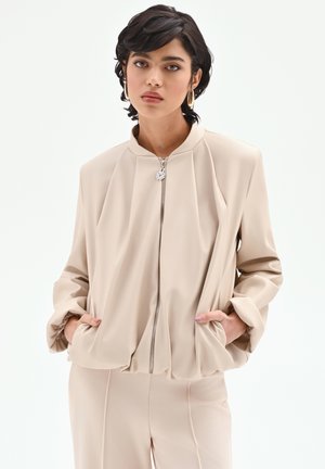Giacca bomber beige con chiusura frontale a zip, design plissettato e maniche lunghe e ampie. Include tasche laterali e una cerniera argentata come dettaglio.